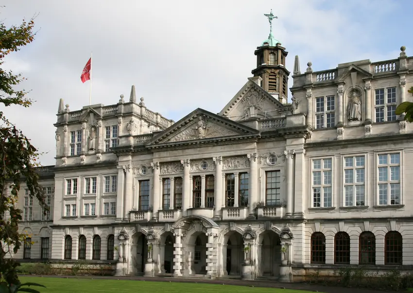 The background is a photograph of a tall, grey, stone building at the University of Cardiff. The front of the building features columns and reliefs, while a red flad flies from the roof.